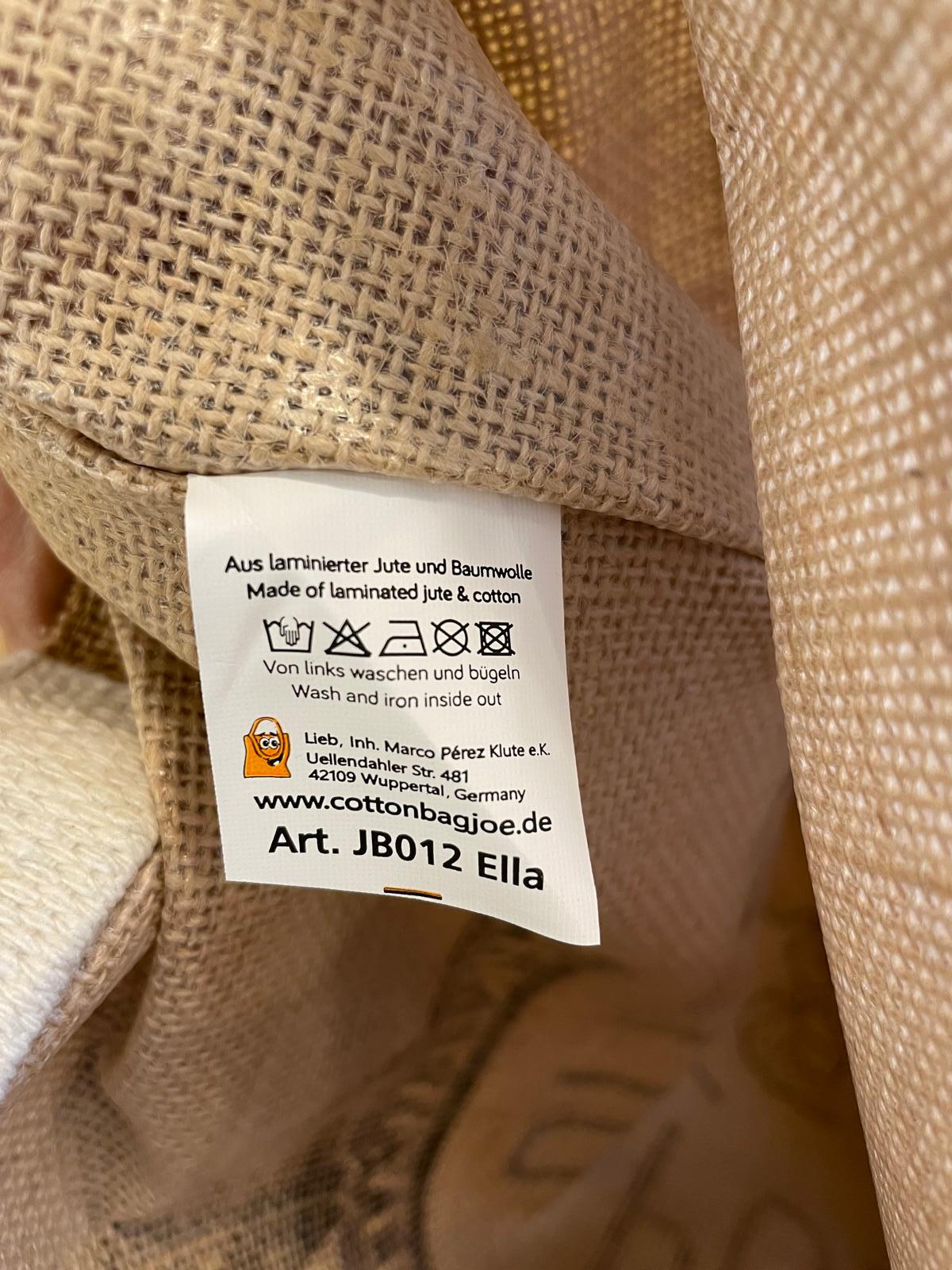 Nahaufnahme eines Etiketts in einer Spreewald Jutetasche, das Pflegehinweise und Herstellerangaben zeigt. Das Etikett gibt an, dass die Tasche aus laminiertem Jute und Baumwolle besteht, mit Waschhinweisen und der Website des Herstellers.